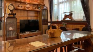 A dining area with a glass table, wooden chairs, a wooden cabinet with shelves, a television, and a giraffe statue on the windowsill. - A Xin B&B (Jinning)