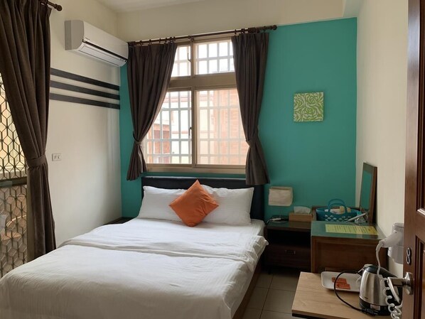Una cama bien tendida con sábanas blancas y un almohadón naranja, una mesita de noche con lámpara y teléfono, y una ventana con cortinas.
