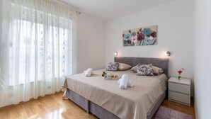 A bedroom with a bed, a nightstand, a painting, and a window with sheer curtains. - Urban Villa Penthouse (Podstrana)