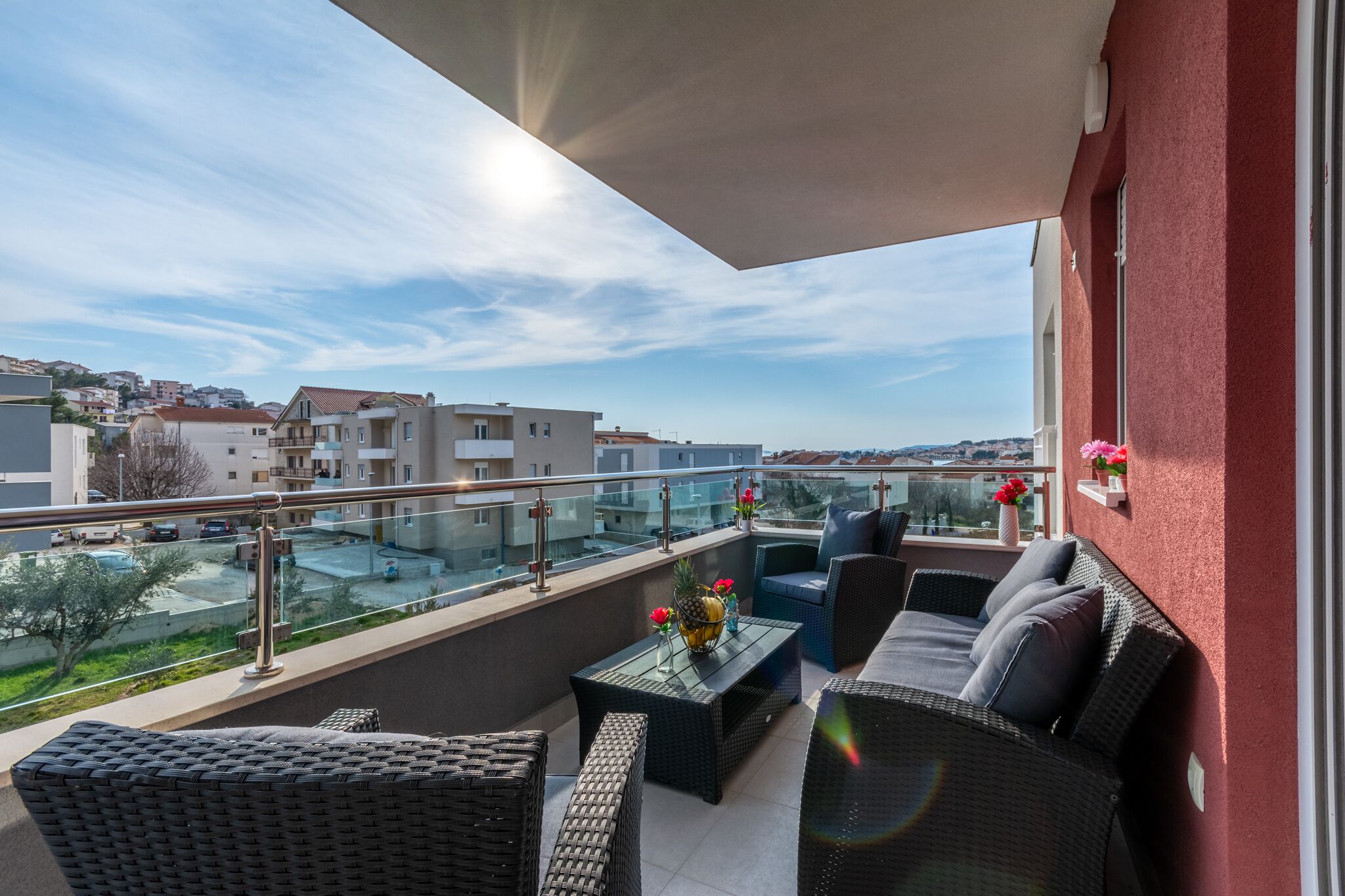 A balcony with wicker furniture, a glass table, and a view of a residential area.
