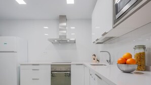Une cuisine moderne avec des meubles blancs, une hotte en acier inoxydable, un four encastré, un évier et un saladier rempli d’oranges posé sur le plan de travail.