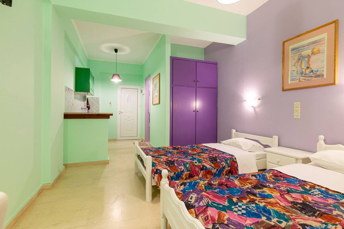 Ein Zimmer mit zwei Betten, einem violetten Kleiderschrank, einer kleinen Küchenzeile und einer Tür zu einem weiteren Raum.