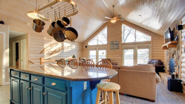 Interior de una cabaña de troncos con una isla de cocina, sillas de madera para comedor, un sofá y un televisor montado en la pared.
