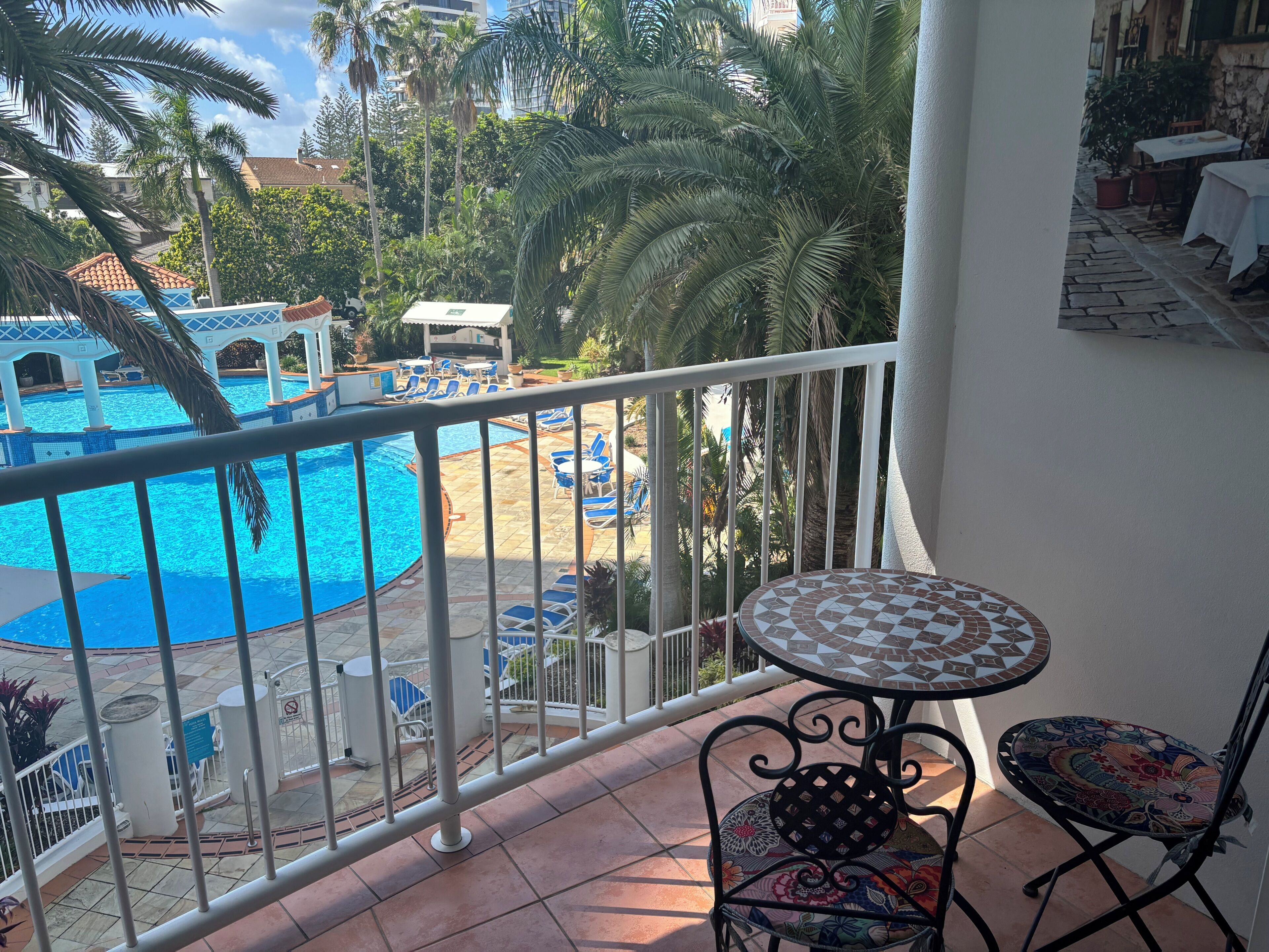 A balcony with a view of a swimming pool, lounge chairs, and a tiled table.