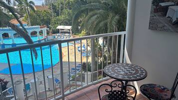 A balcony with a view of a swimming pool, lounge chairs, and a tiled table.