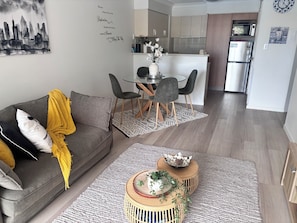 A modern living room with a grey sofa, a dining table with chairs, a kitchen area, and a refrigerator. - Charming Broadbeach unit - Soothing pool views (Broadbeach)