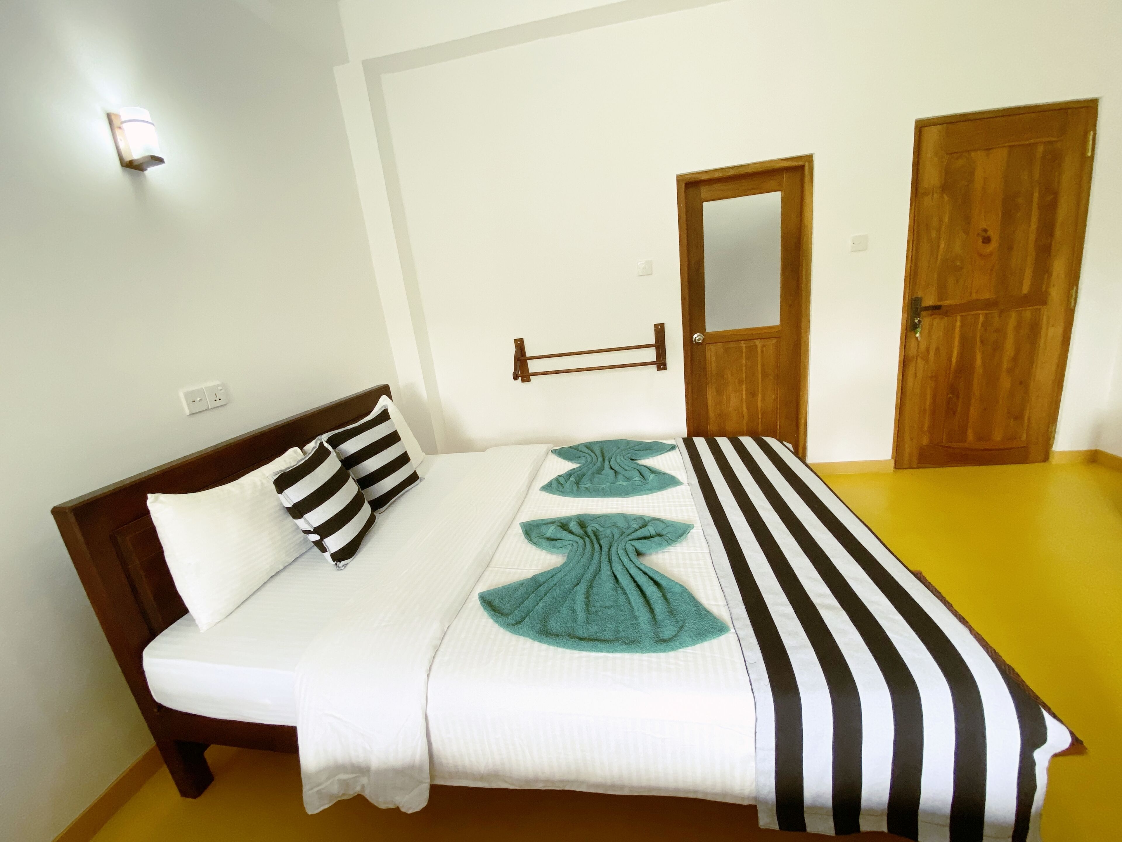 A neatly made bed with striped bedding and a teal towel, a wooden door, and a wall-mounted light fixture.