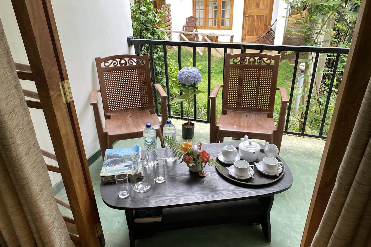 Un balcón con dos sillas de madera, una mesa dispuesta con tazas de té, platitos y una tetera, y una bandeja con botellas de agua.