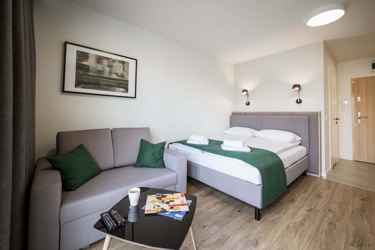 A modern hotel room with a grey sofa, a bed with green bedding, a coffee table with remote controls, and a framed picture on the wall.