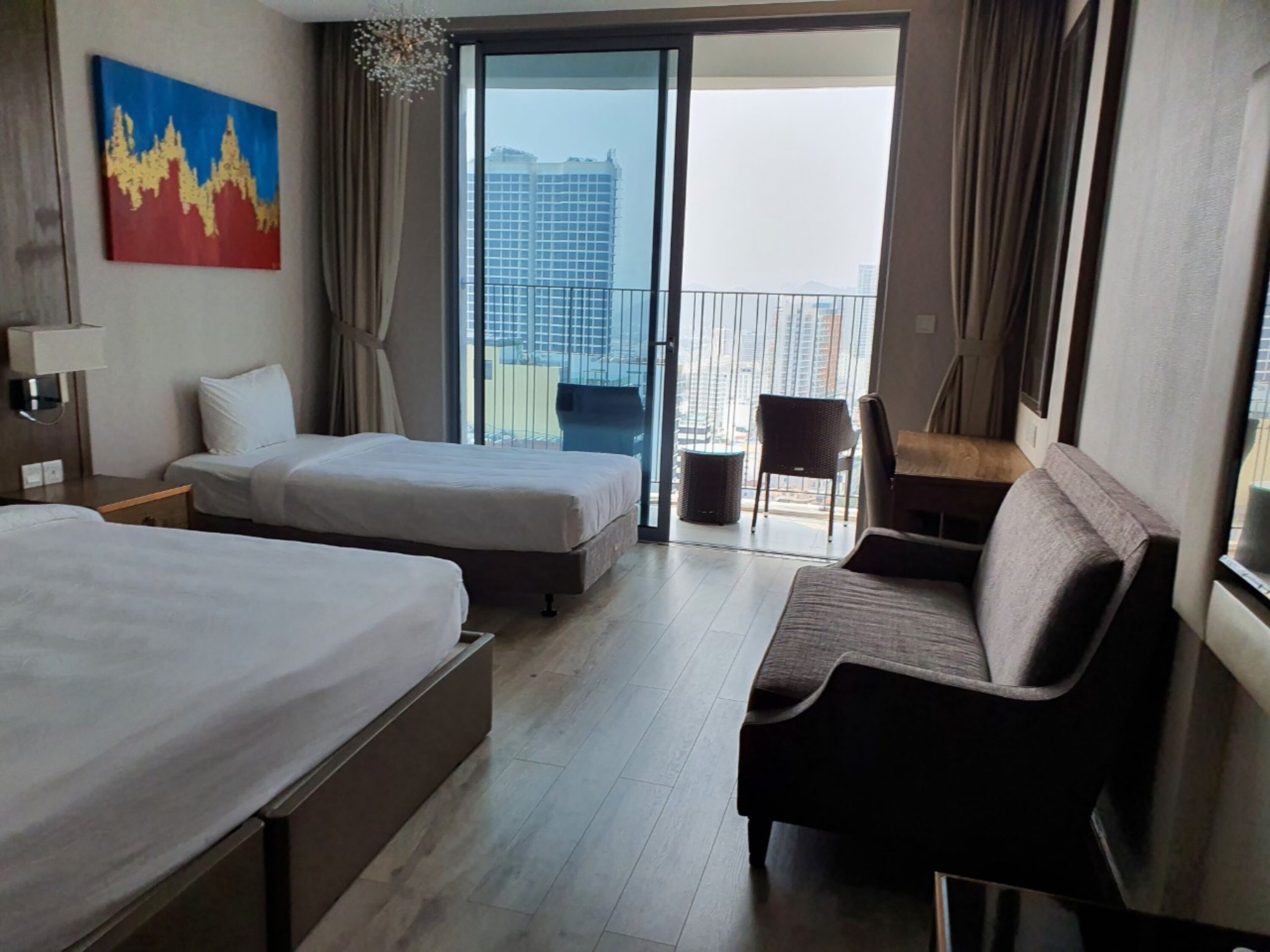 A hotel room with two beds, a sofa, a desk, and a balcony with a city view.