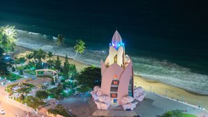 A night view of a coastal area with a prominent, illuminated structure resembling a rocket, surrounded by a landscaped park with palm trees and a beach.