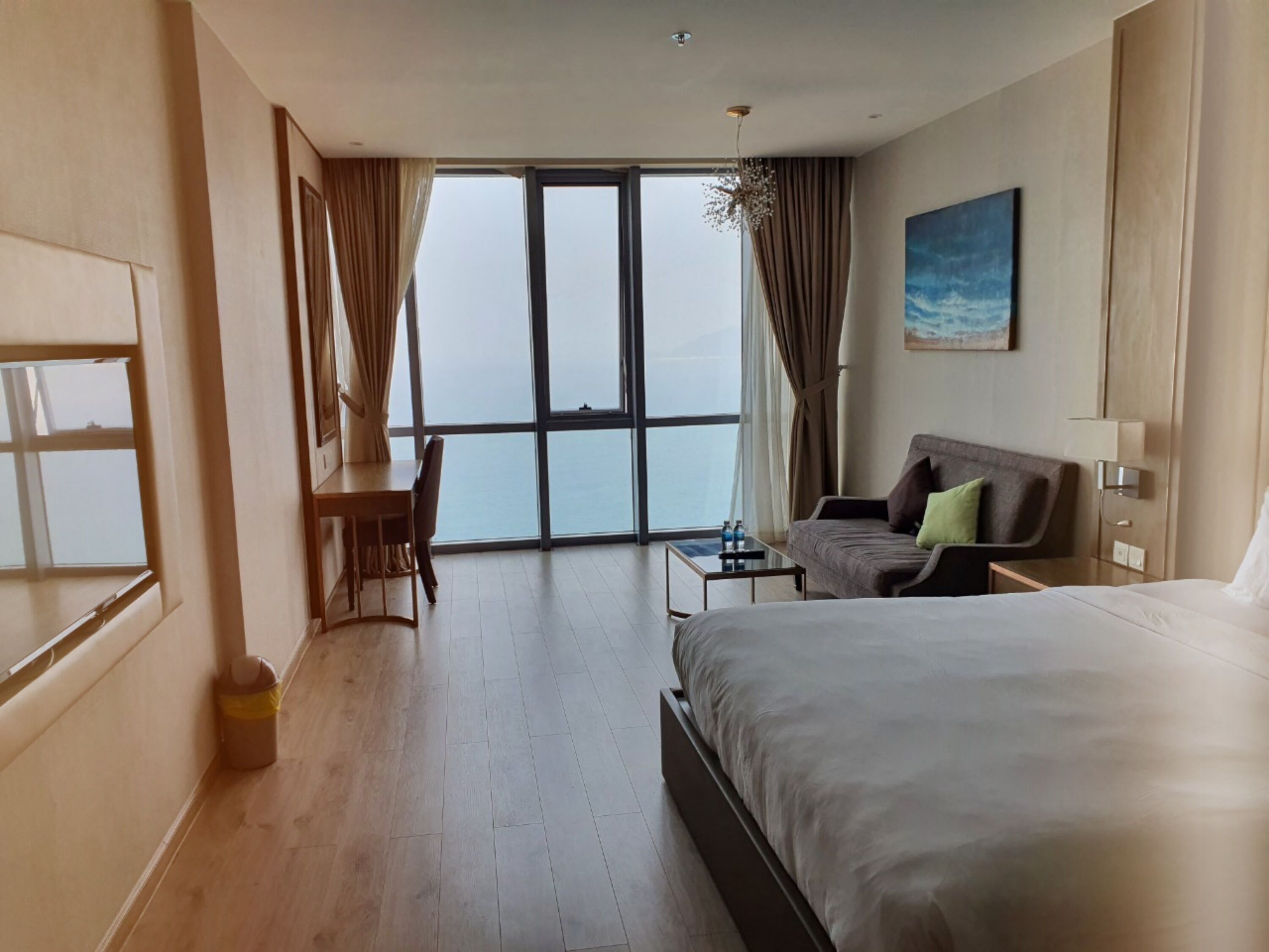 A hotel room with a large bed, a desk, a chair, and a view of the ocean through a large window.