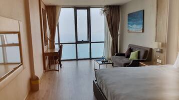 A hotel room with a large bed, a desk, a chair, and a view of the ocean through a large window.