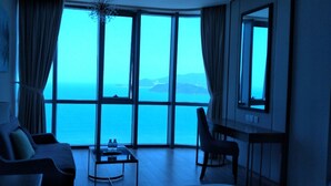 A room with a large window offering a view of the sea and distant islands. There is a sofa, a chair, and a small table with two bottles on it.