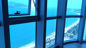 A high-rise hotel room with large windows offering a view of a coastal area, including a beach and a cityscape.