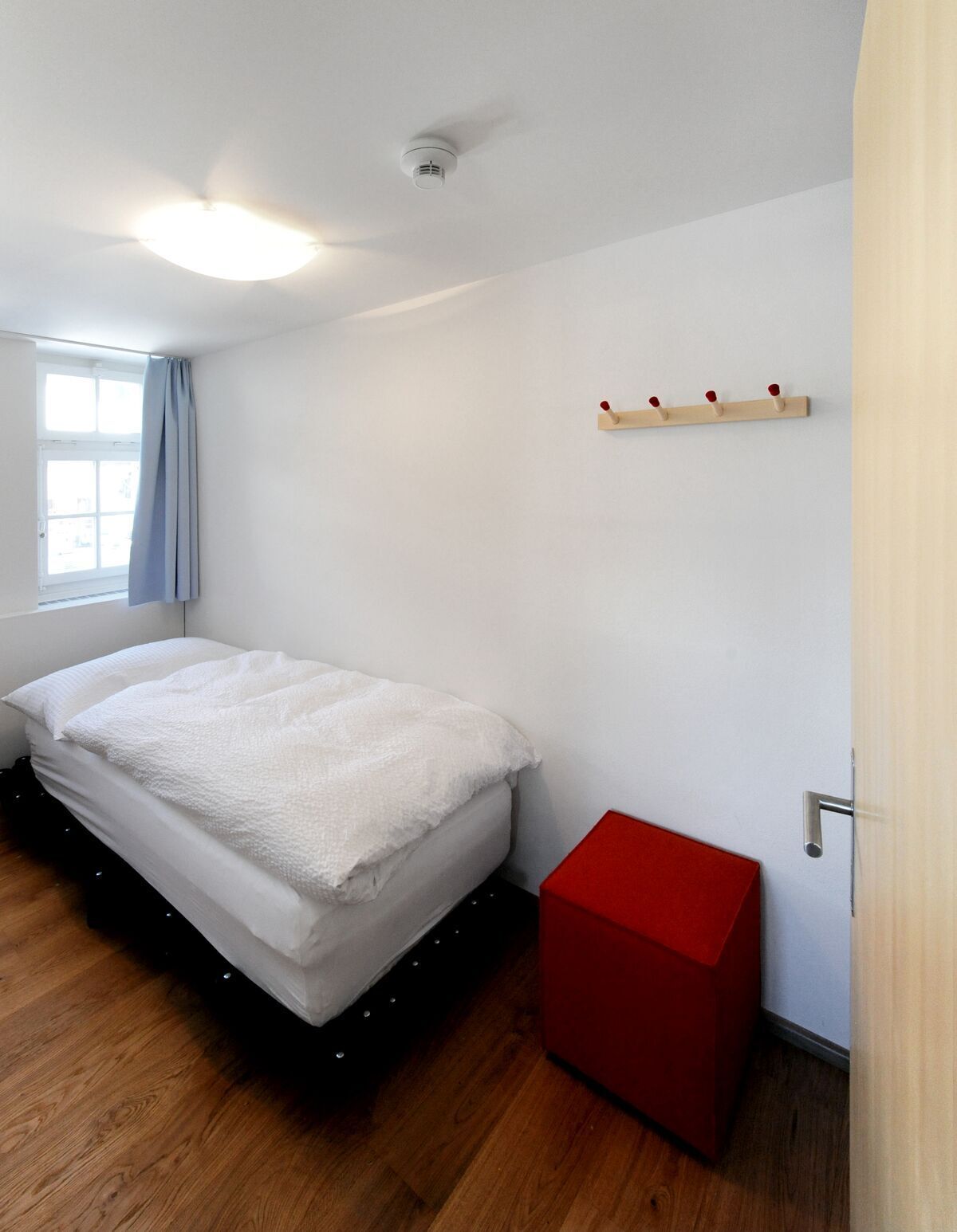 Un dormitorio con una cama, un baúl rojo, un estante de madera con ganchos y una ventana con cortinas.