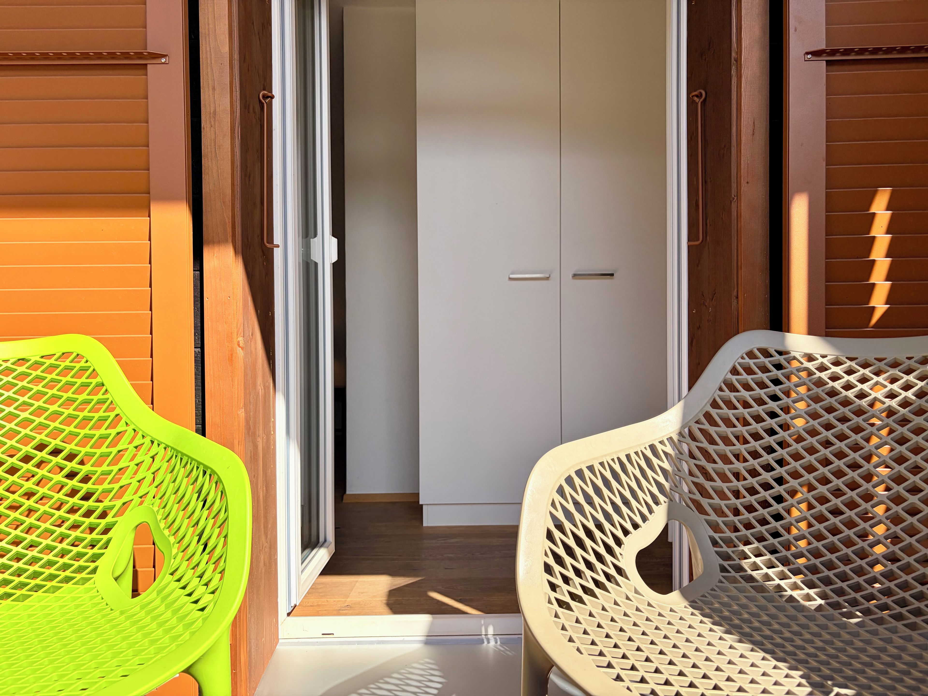 Una habitación con puerta corredera que da a un pasillo, una puerta blanca y dos sillas con diseño de celosía.