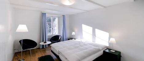 A modern bedroom with a large bed, two bedside tables, a chair, a small table, and a window with curtains.