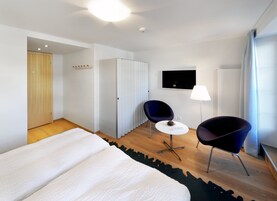 A modern hotel room with a large bed, two chairs, a small table, a TV, and a wardrobe.