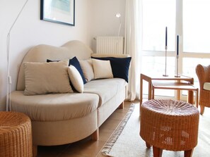 Ein gemütliches Wohnzimmer mit einer Couch, zwei Beistelltischen, einem Rattanasitzhocker und Blick ins Freie durch ein großes Fenster.