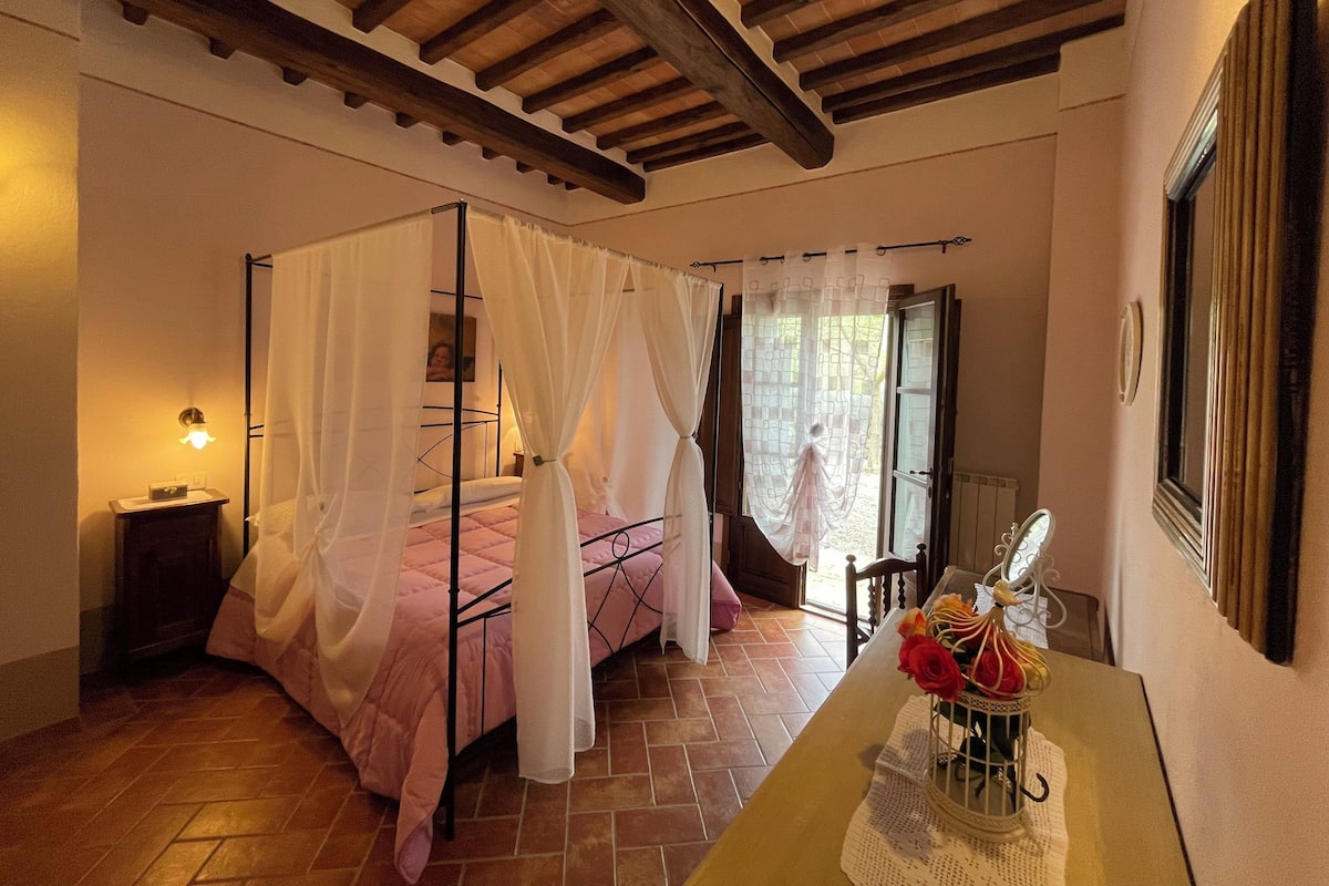 Un dormitorio con cama con dosel, una mesita de noche, cortinas en la ventana y vistas al exterior.