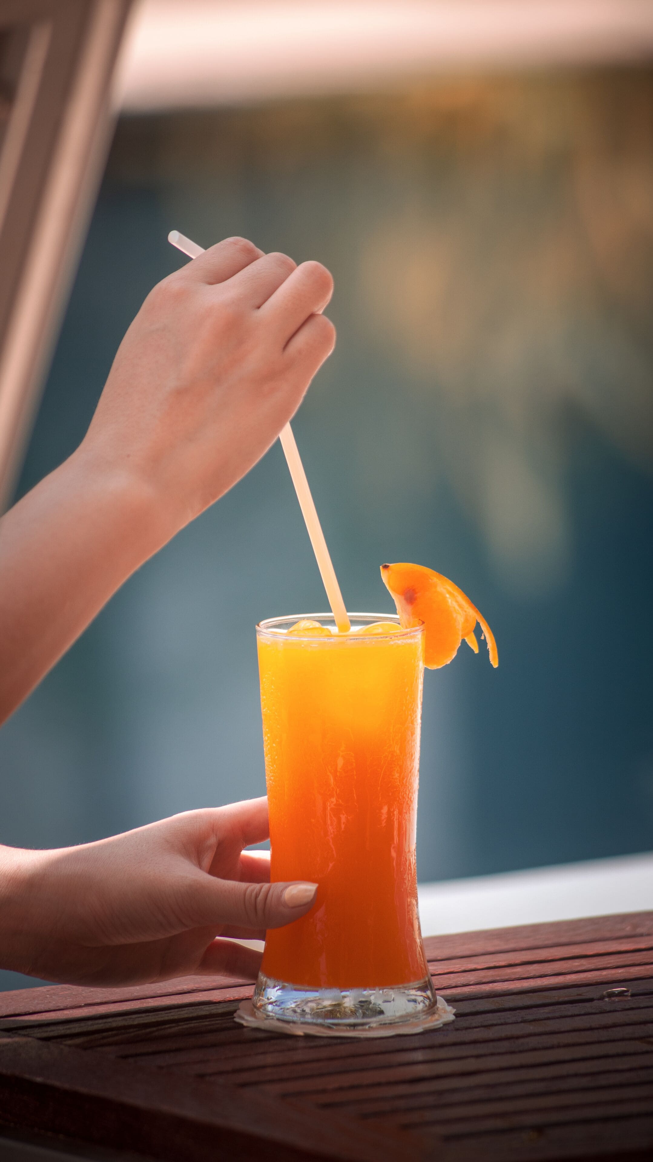 A hand holding a glass with a straw, containing a bright orange beverage, garnished with an orange slice, on a wooden table.