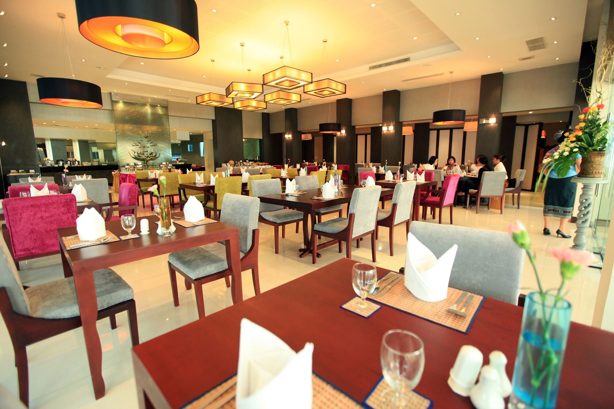 A hotel dining area with multiple tables and chairs, a bar in the background, and a modern chandelier.