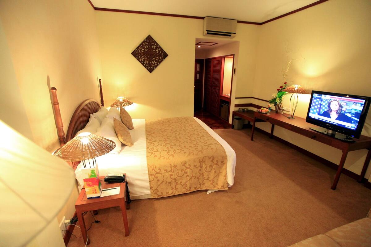 A hotel room with a bed, bedside table, lamp, TV, and a desk with a chair.
