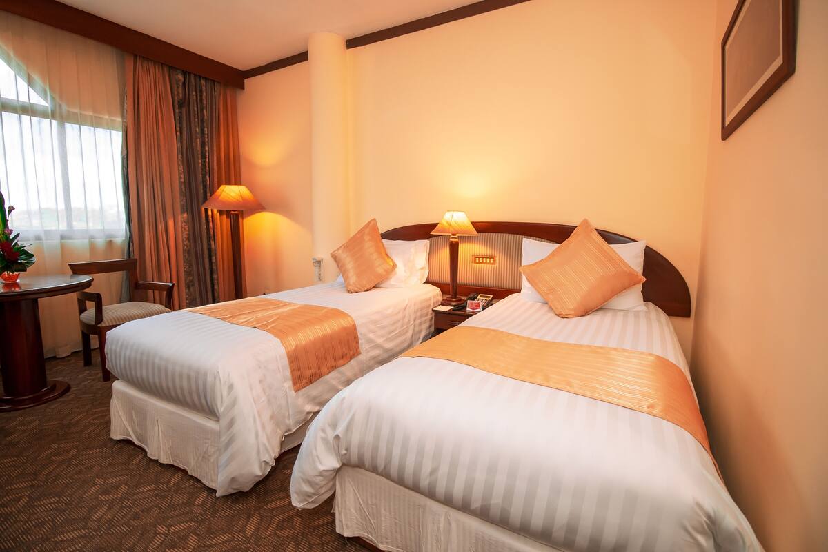 A hotel room with two beds, a desk, a chair, a lamp, and a window with curtains.