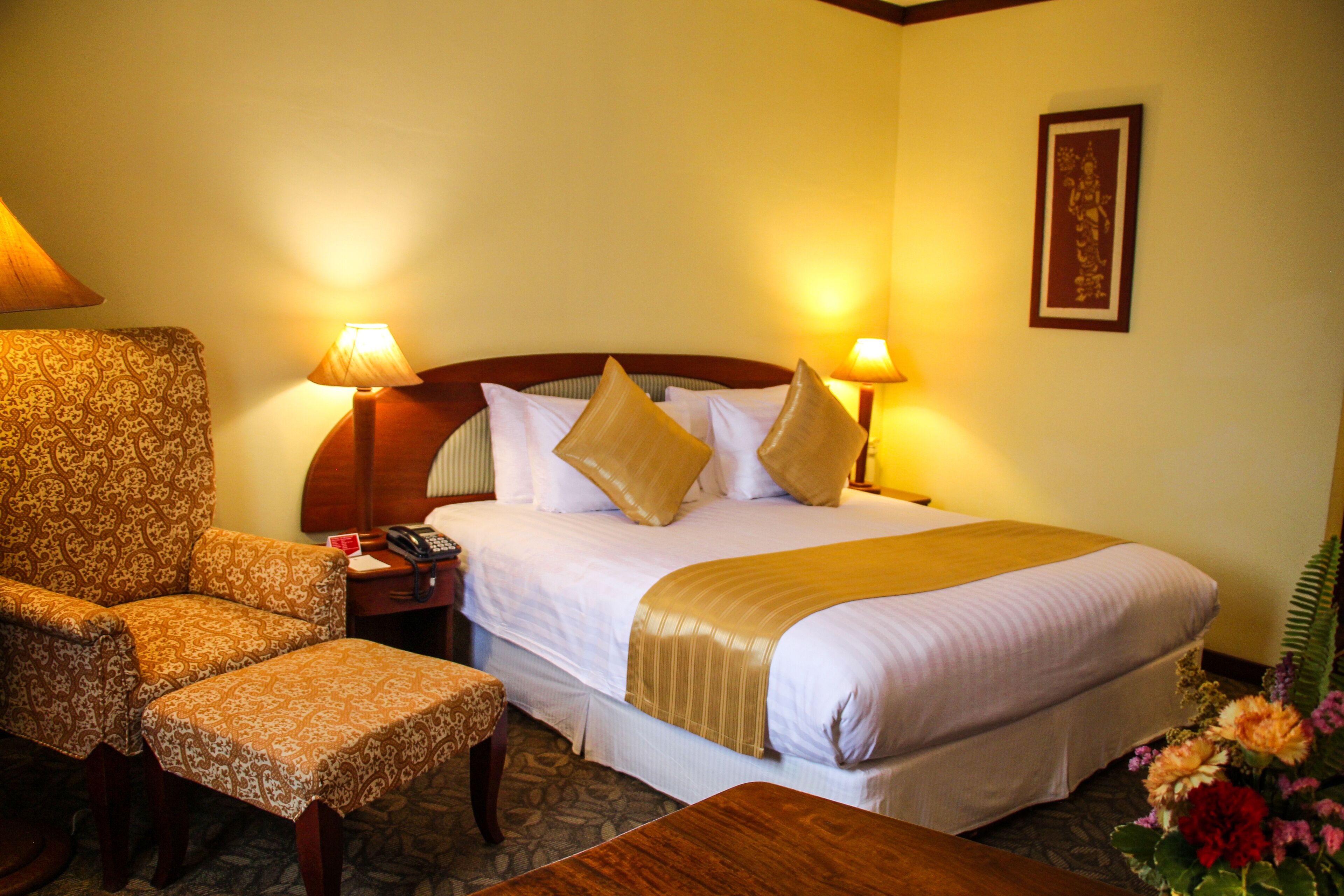 A hotel room with a bed, two bedside lamps, a chair, a table, and a framed picture on the wall.