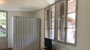 Une pièce avec un plancher en bois, une porte coulissante, une fenêtre avec des stores, une commode en bois sur laquelle se trouve un téléviseur, et un radiateur.