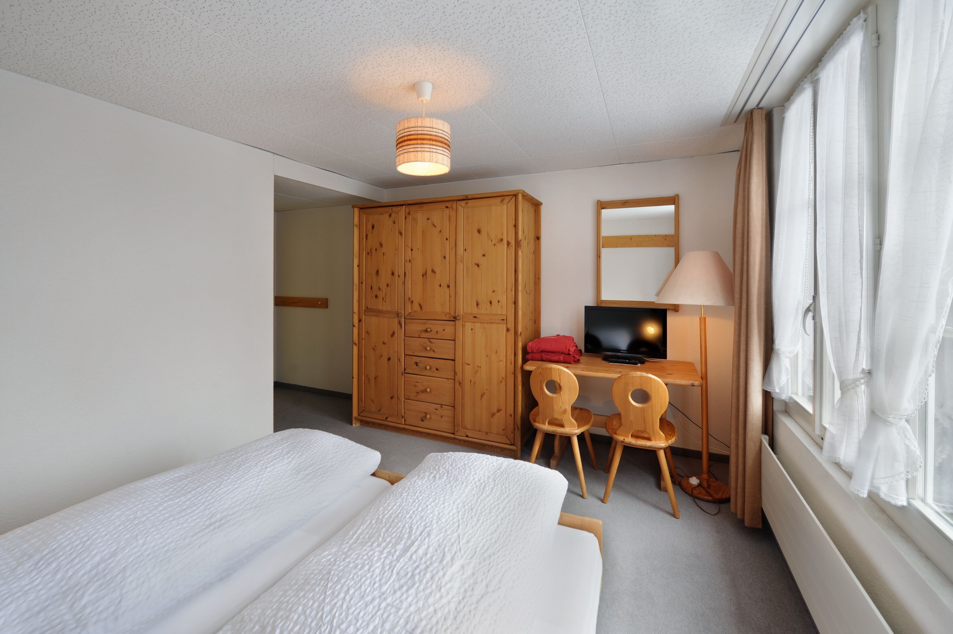 A hotel room with a bed, a desk with a computer, a wardrobe, and a window with curtains.