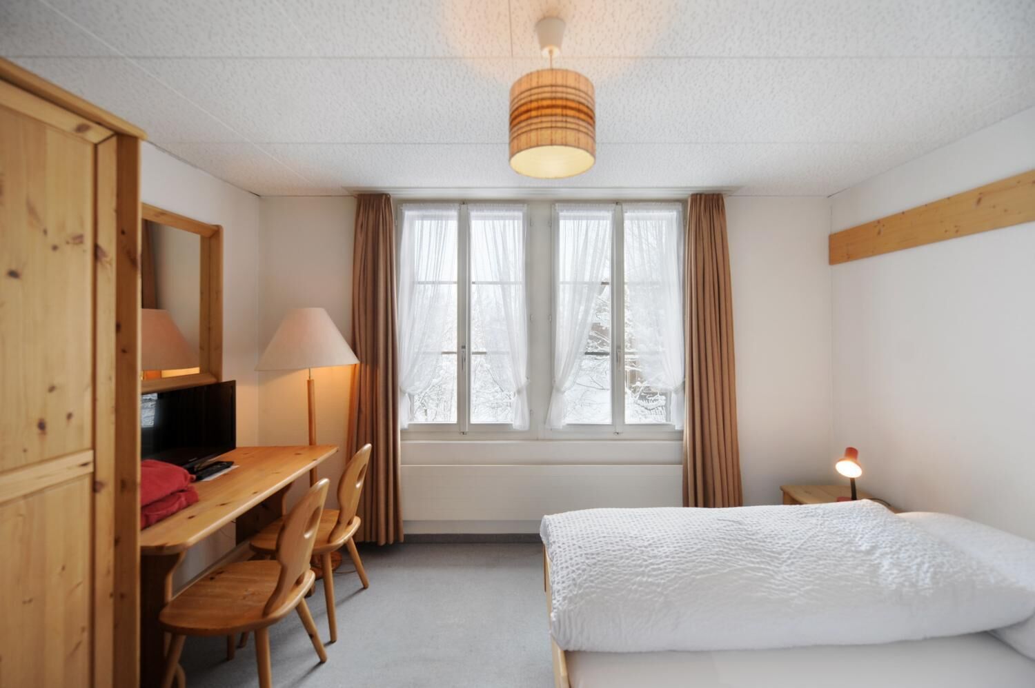 A room with a bed, a desk with a chair, a lamp, and a window with curtains.