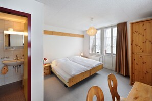 A bedroom with a wooden bed, a bedside table, a nightstand, a wooden wardrobe, a window with curtains, and a bathroom with a sink and mirror. - Hotel Sternen Budget  (Lenk)