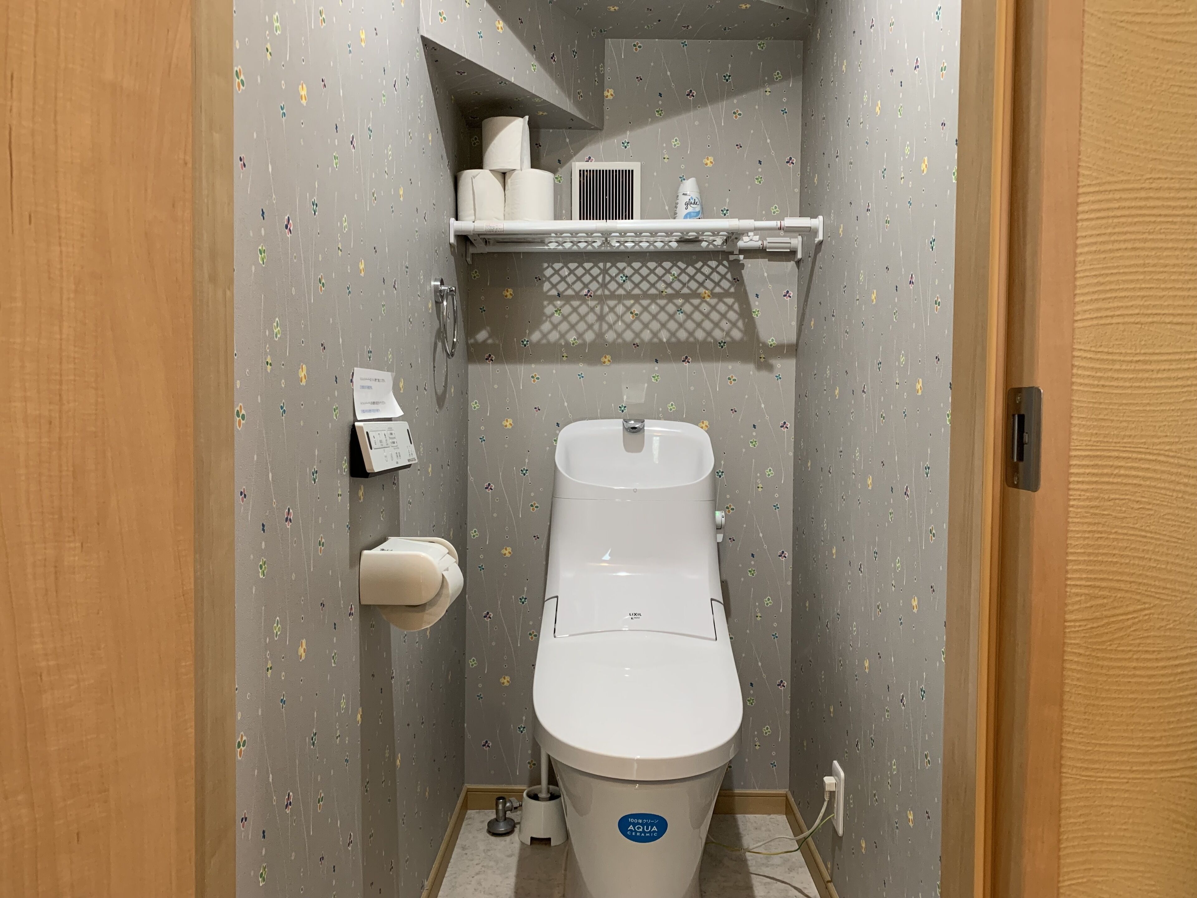 Un baño pequeño con un inodoro, dispensador de toallas de papel y un estante con papel higiénico y una botella.