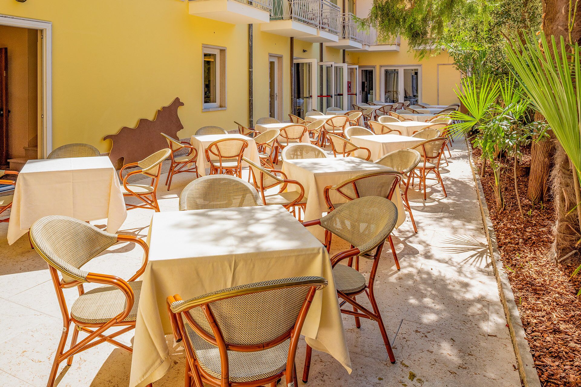 An outdoor dining area with wicker chairs and tables, surrounded by a yellow building with balconies.