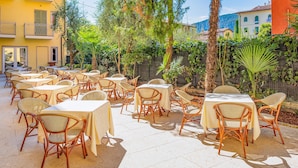 An outdoor patio with tables and wicker chairs, surrounded by greenery and buildings. - Hotel Centrale (Garda)