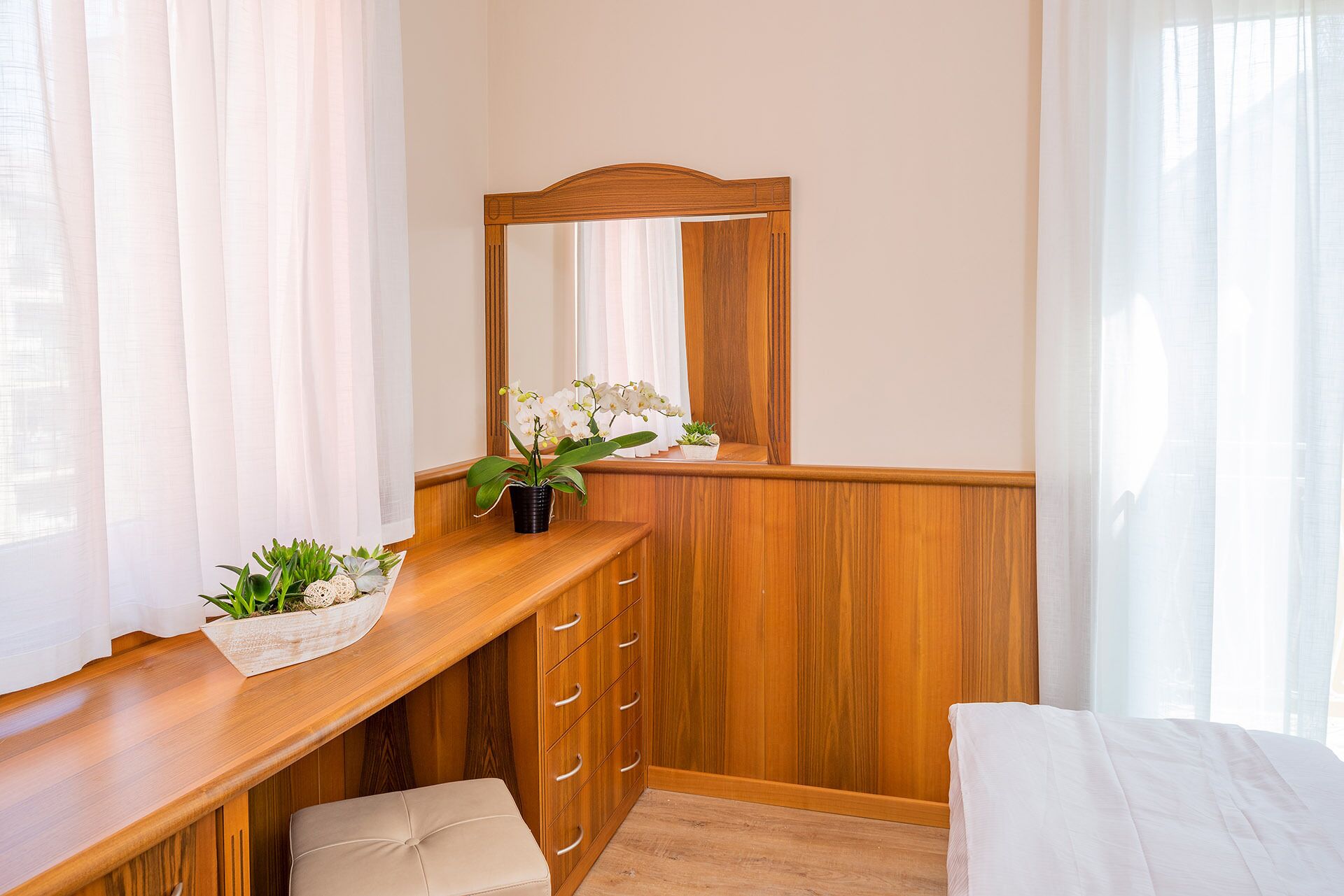 A bedroom with a wooden dresser, a mirror, a bed, and a window with sheer curtains.