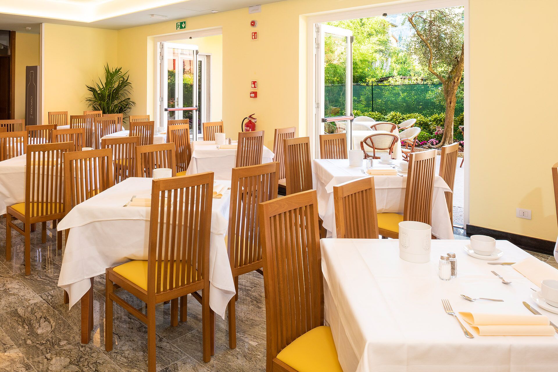 A dining area with multiple tables and chairs, a fire extinguisher, and a view of the garden.