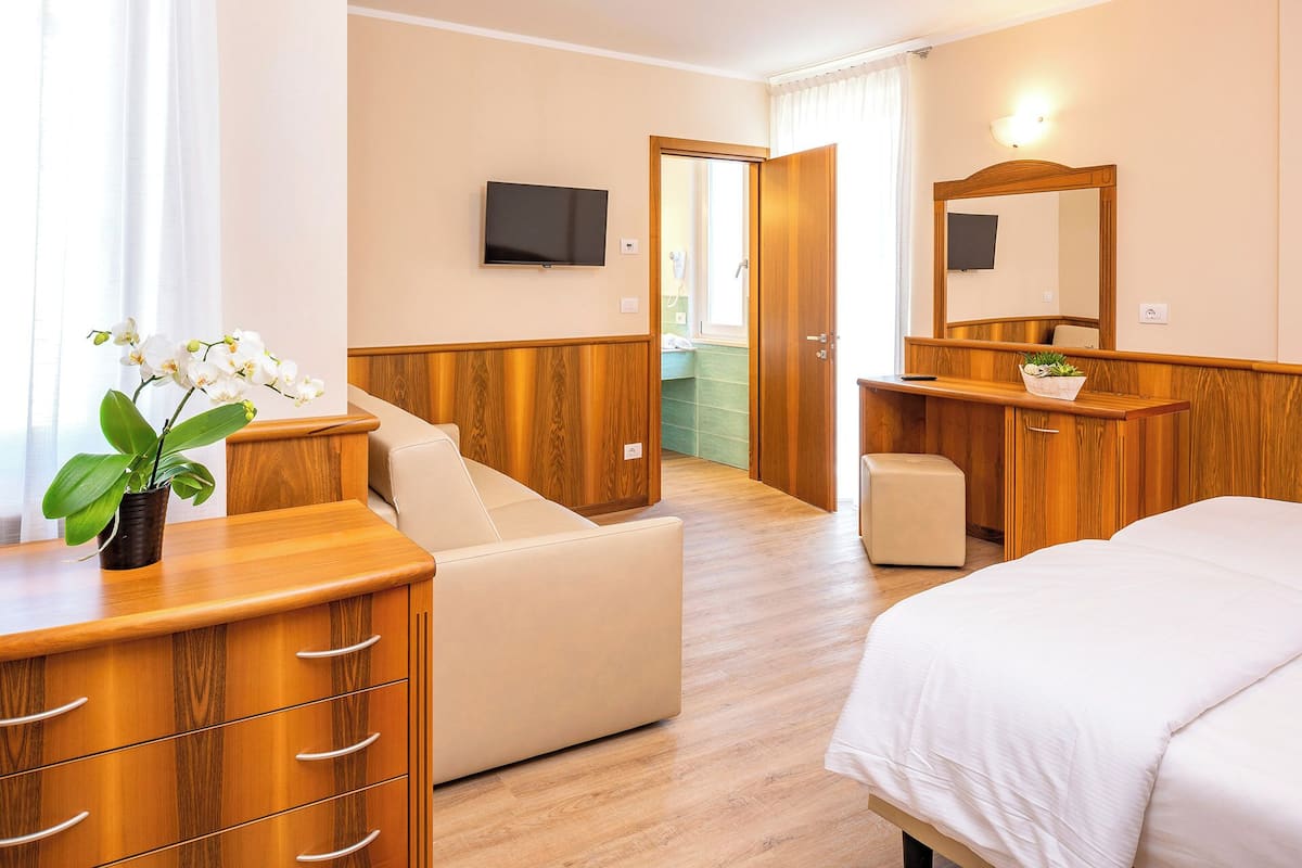 A hotel room with a bed, a wooden dresser, a small table, a chair, a TV, and a bathroom visible through an open door.