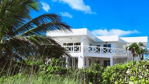 Un bâtiment blanc à deux étages, doté d’un balcon, entouré d’une végétation luxuriante et de palmiers.