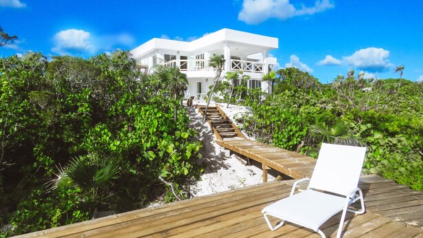 Une terrasse en bois m ène à un bâtiment blanc doté d’un balcon, le tout étant entouré d’une végétation luxuriante.