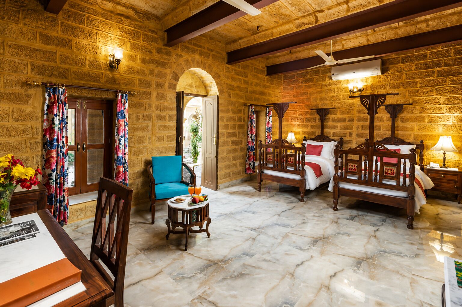 Una habitación con paredes de piedra, una cama de madera, una silla azul, una mesita con fruta y una puerta que da al exterior.