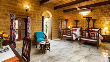 Una habitación con paredes de piedra, una cama de madera, una silla azul, una mesita con fruta y una puerta que da al exterior.