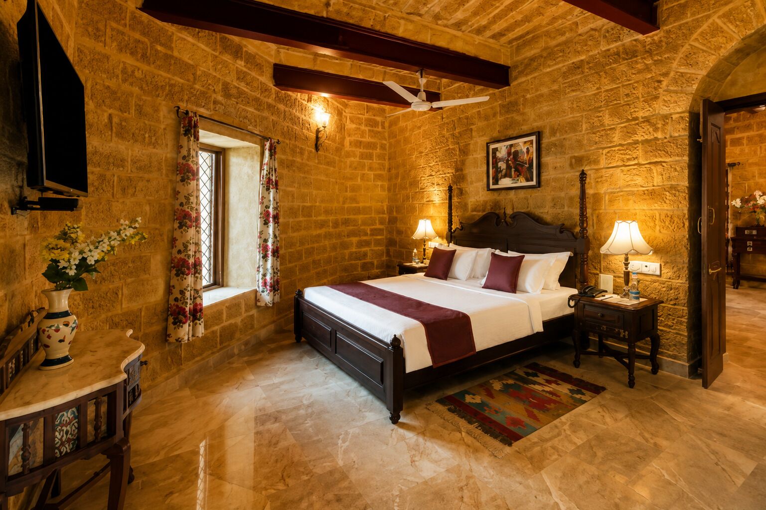 Un dormitorio con pared de piedra, cama con cabecera de madera, mesita de noche con lámpara y ventana con cortinas florales.