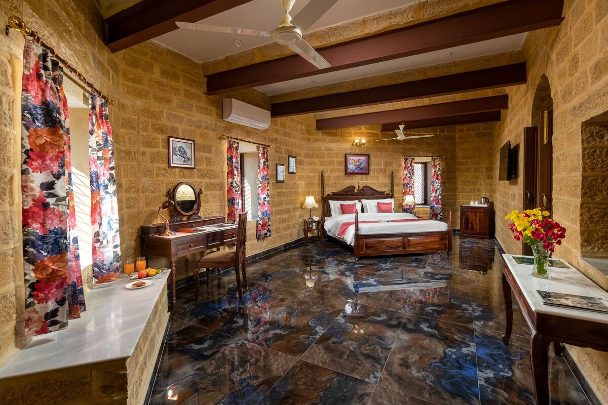 Um quarto com cama, escrivaninha, cadeira, mesa com um vaso de flores e ventoinha no teto.