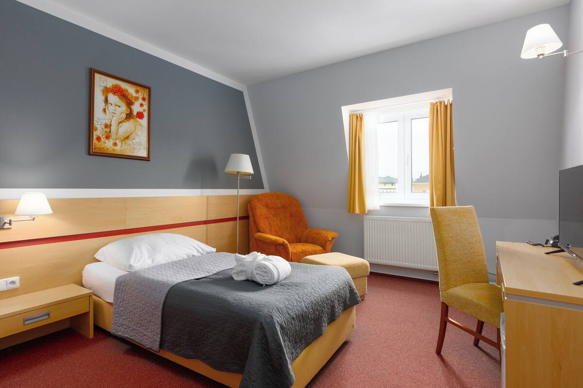 Une chambre d’hôtel avec un lit, un bureau, une chaise, une lampe et une fenêtre avec des rideaux.