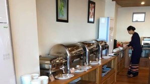 Un buffet avec plusieurs présentoirs de nourriture, une personne qui sert les plats et un espace cuisine en arrière-plan.