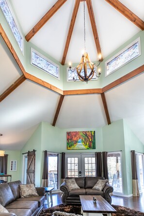 A spacious living room with a high ceiling, wooden beams, and a chandelier. There is a large painting on the wall and a view through the glass doors.