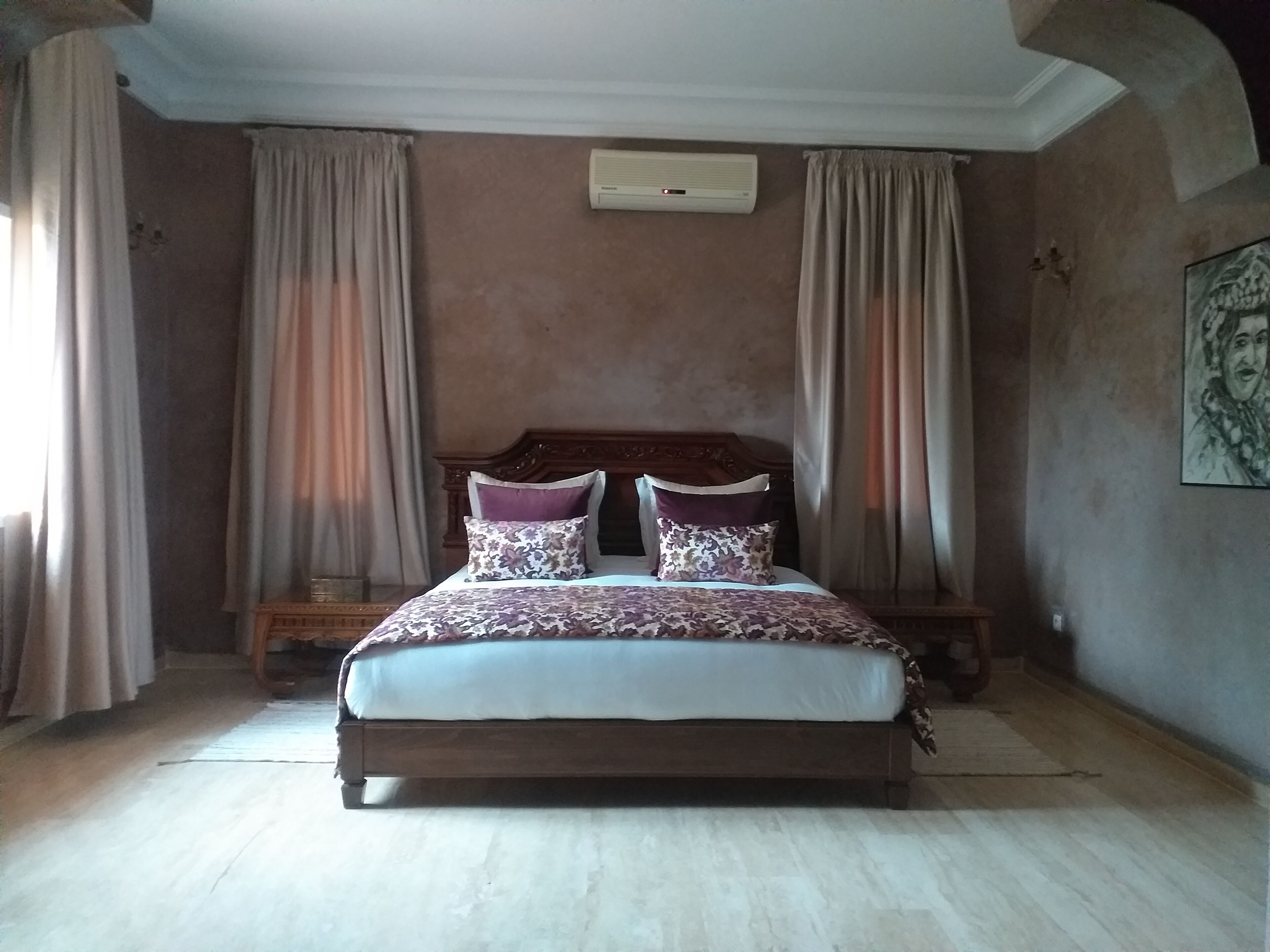A bedroom with a large bed, curtains, a wall-mounted air conditioner, and a framed picture on the wall.